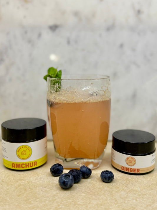 An orange-colored drink with a mint sprig, behind loose blueberries, and next to jars of Amchur and Ginger.