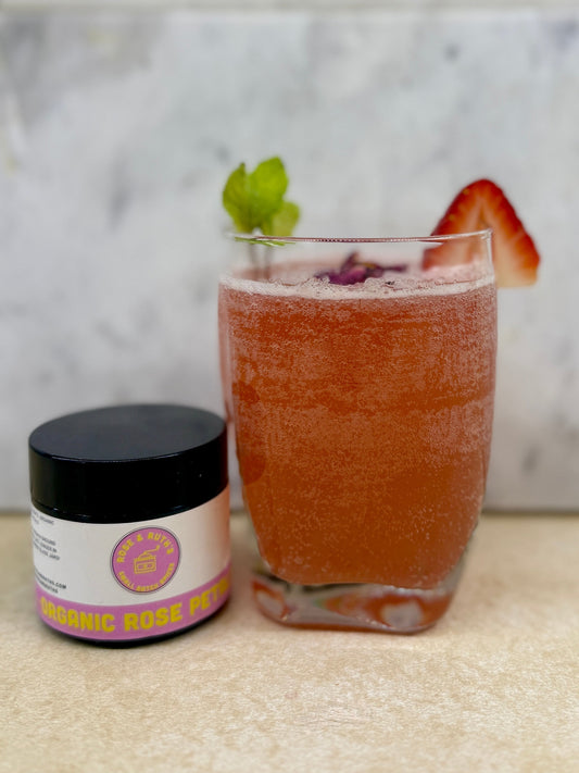 A pink, bubbly beverage in a clear glass with a strawberry, mint, and rose petal garnish, next to a jar of Organic Rose Petals.