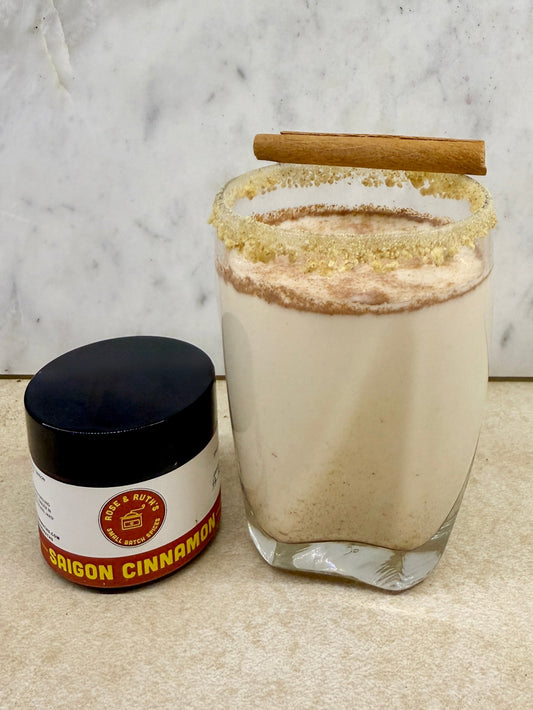 Milky drink topped with ground Saigon Cinnamon, graham cracker-rimmed glass, and cinnamon stick on top next to a jar of Saigon Cinnamon.