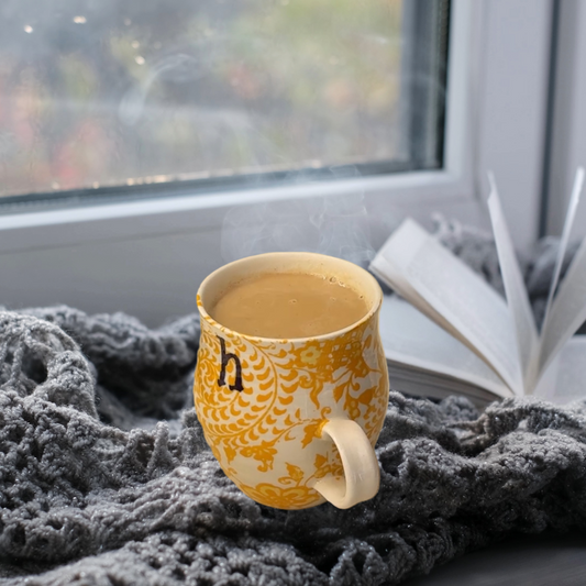 A yellow and white patterned mug of steaming chai on top of a soft gray blanket next to a book by a window.